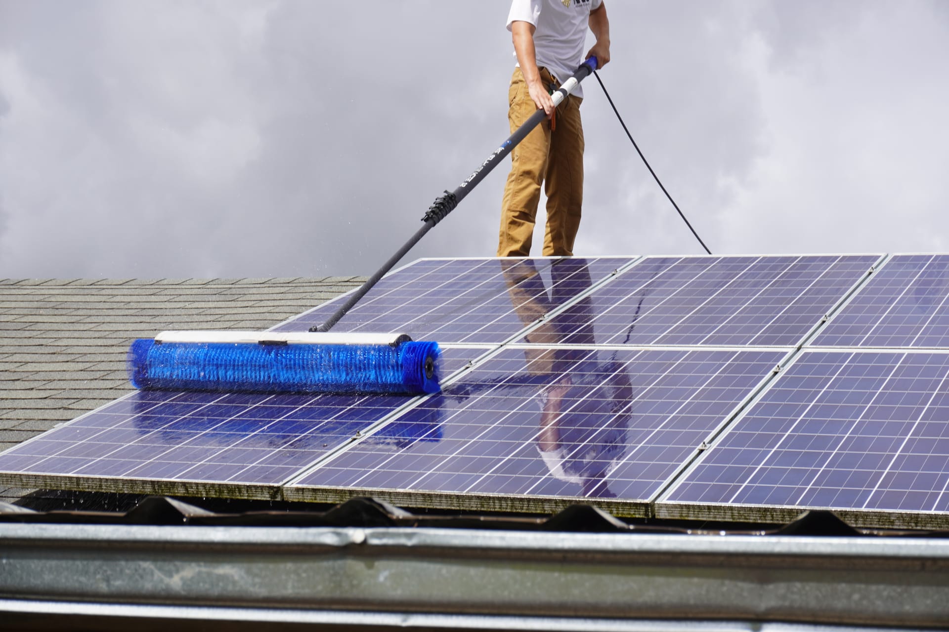A technician washing rooftop solar panels with a blue rotary brush, panels wet and reflective